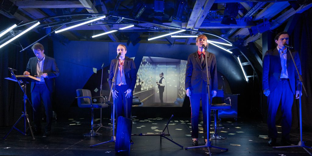 Vier Menschen stehen nebeneinander auf einer blau ausgeleuchteten Bühne. SIe tragen Anzüge, drei von ihnen singen in Mikrofone, einer spielt ein Glockenspiel.