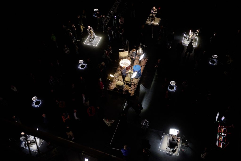 A bird's-eye view of the stage: musicians sit scattered across islands of light on an otherwise black stage. In the center is a larger, illuminated platform on which two performers can be seen.