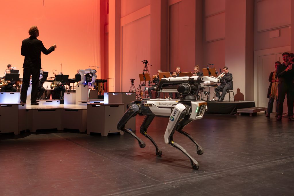 Scene from “Robot Symphony”: a robot in the shape of a dog dances in front of an orchestra seated on platforms.