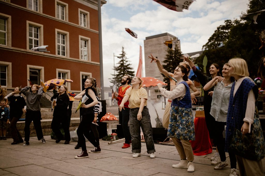 On a rooftop terrace against the backdrop of the city, a group of young people stand laughing and throwing paper airplanes into the air.