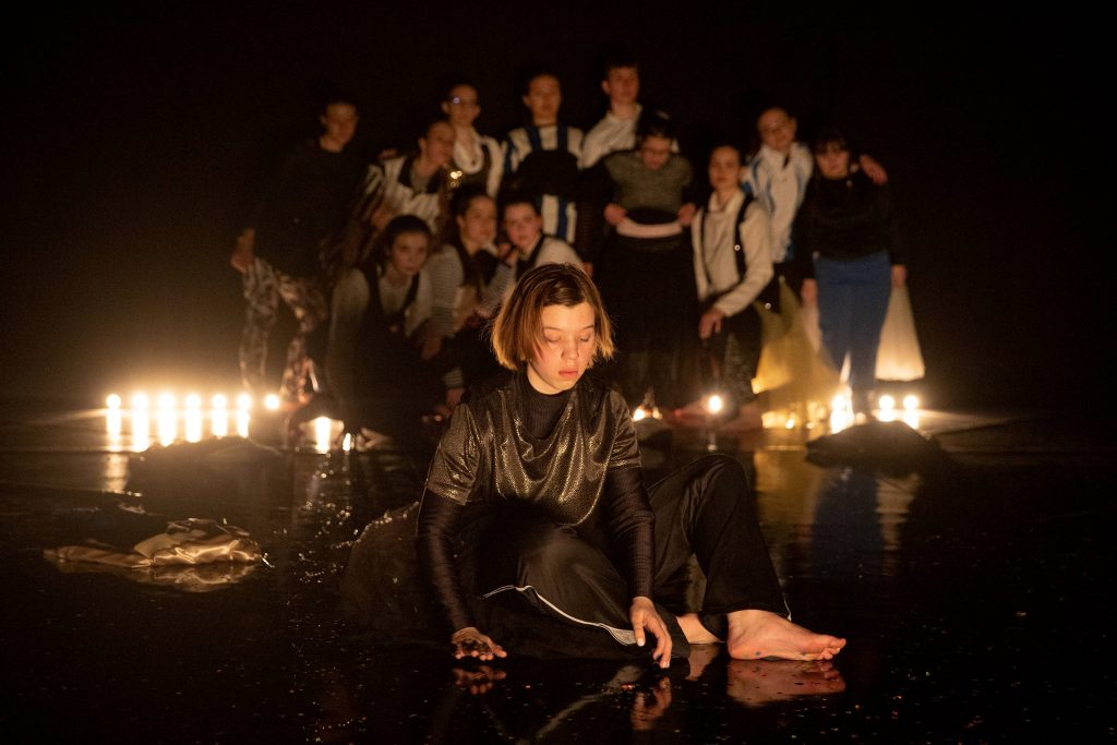 Scene from “FOMO”: a young girl dressed in dark clothes sits alone on the stage floor. A large group of teenagers stare at her from the background.