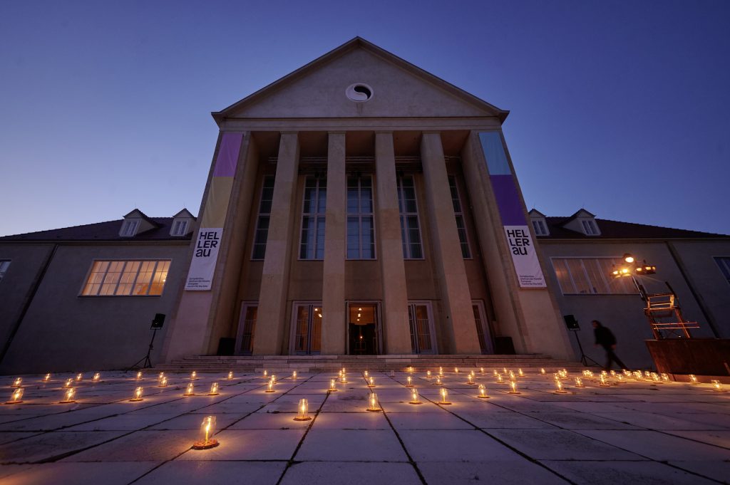 Fassade des Festspielhauses Hellerau vor Abendhimmel. Auf dem gepflasterten Vorplatz steht ein Meer an Lämpchen.