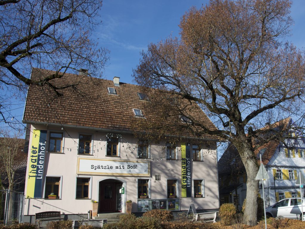 View of the Lindenhof Theater from the outside: a two-story building, with banners bearing the theater's name decorating the left and right sides of the building. Above the entrance hangs a large sign with the inscription “Spätzle mit Soß” (Spätzle with sauce).