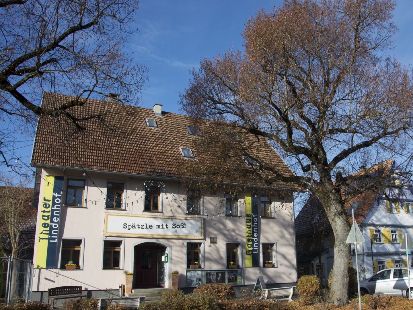 View of the Lindenhof Theater from the outside: a two-story building, with banners bearing the theater's name decorating the left and right sides of the building. Above the entrance hangs a large sign with the inscription “Spätzle mit Soß” (Spätzle with sauce).
