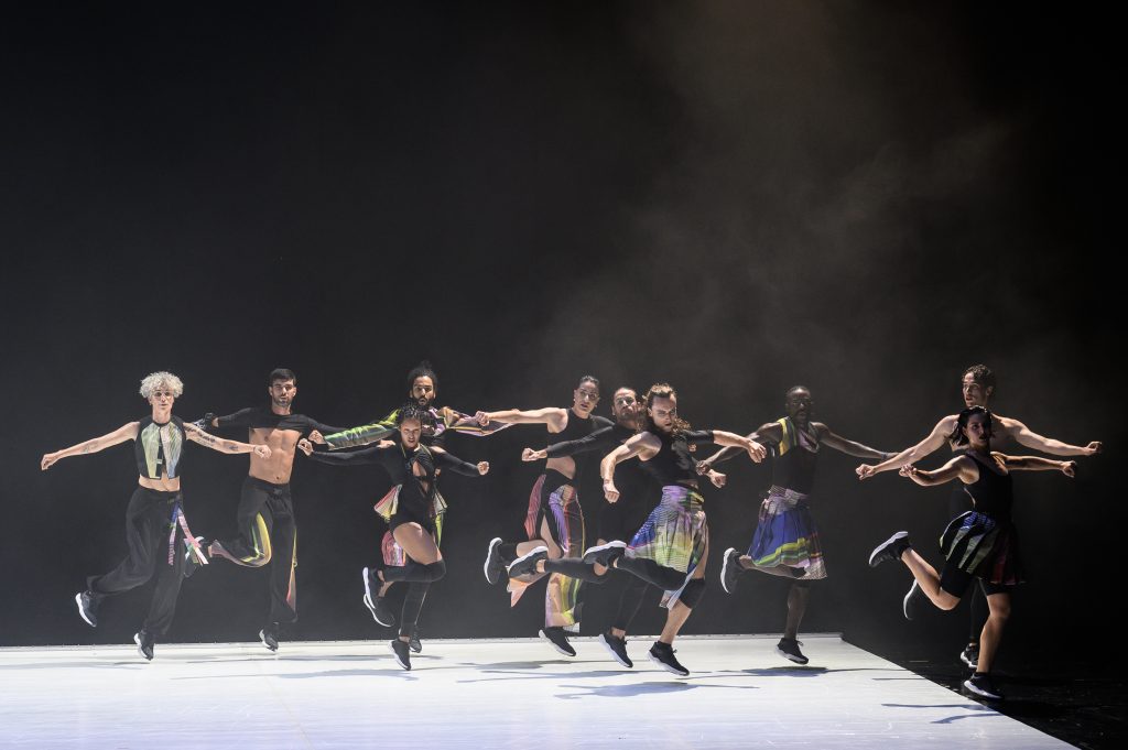 Scene from “Carcaca”: Ten dancers in different costumes dance in two rows in front of a black background with their arms stretched out to the sides.