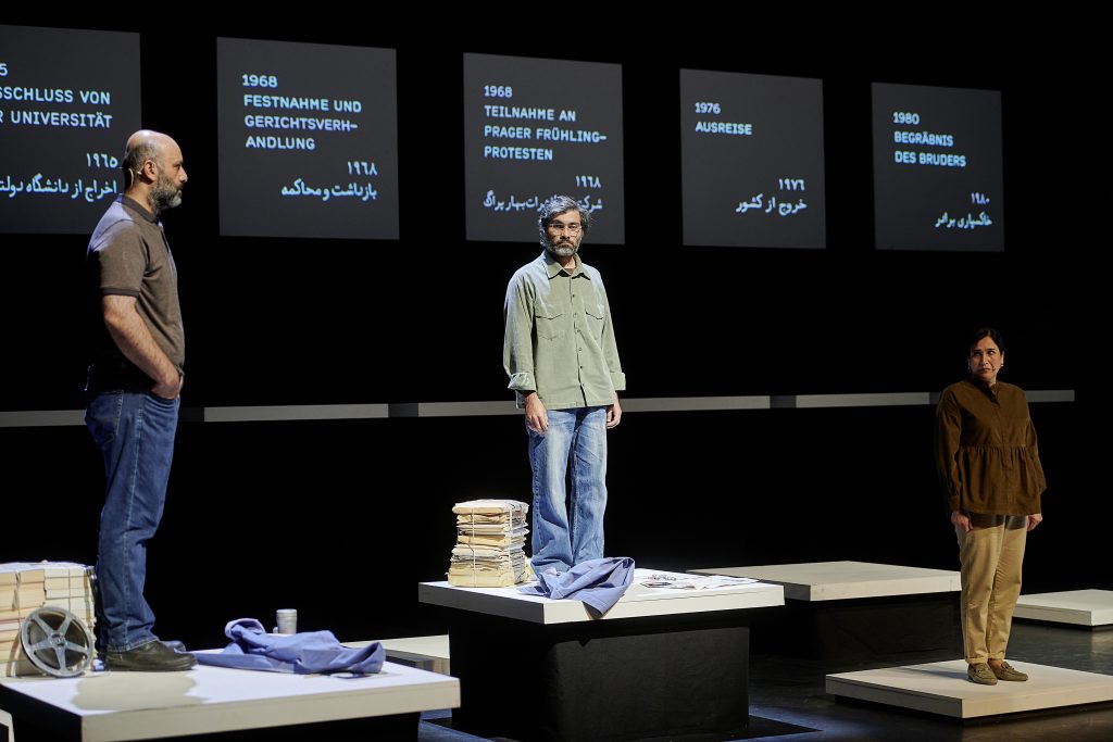 Scene from “Tentative Title: Brasch”: Two men and a woman stand on different platforms, facing each other. Stacks of paper and books are next to them. In the background, a timeline with historical events is projected.