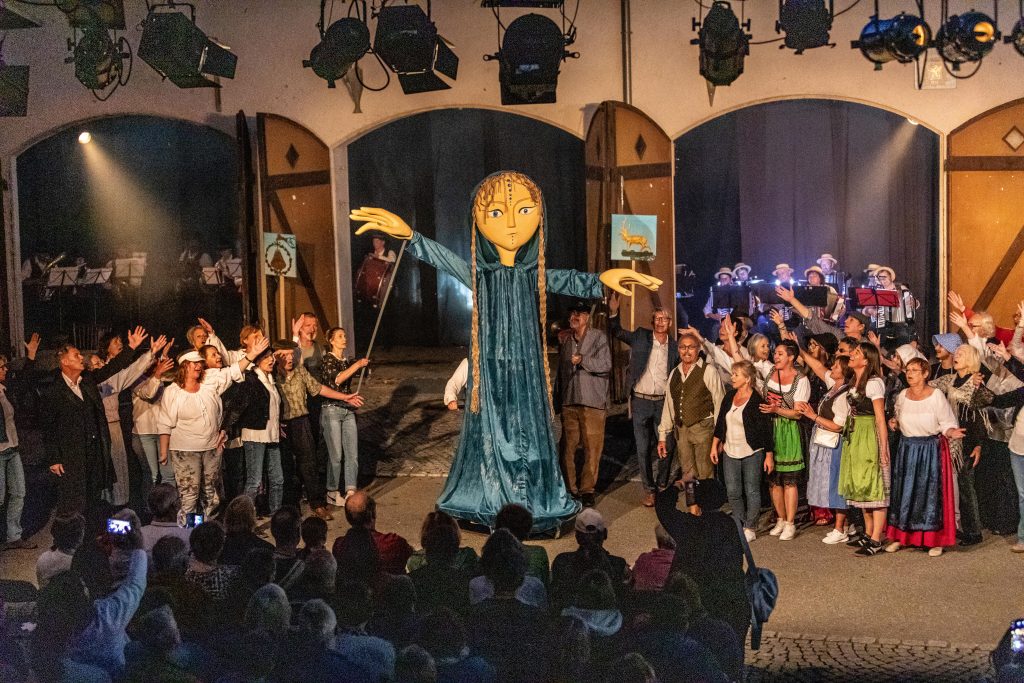 An oversized marionette can be seen on an open stage under the open sky. Groups of people stand to the left and right of it, pointing at it and singing. In the background, against the backdrop of a house with large, open doors, an orchestra can be seen.
