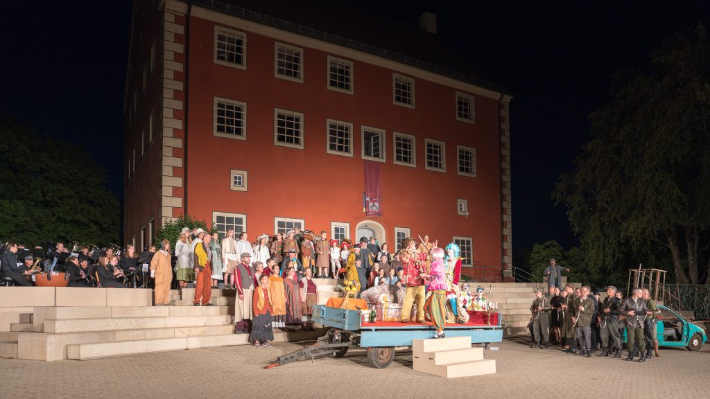 Scene from a play performed in an open-air market square: People of all ages stand together in groups wearing different costumes. An open trailer serves as the stage. In the background, a children's choir and an orchestra can be seen against the backdrop of a red house.
