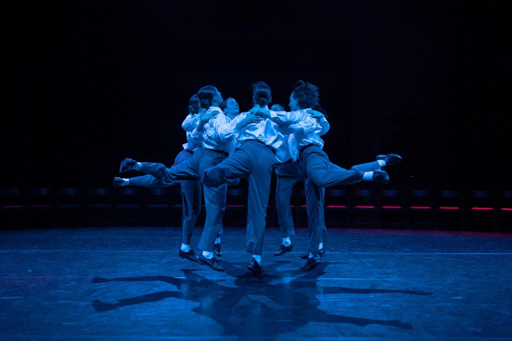 Scene from “Ring”: Bathed in blue light, dancers form a circle, supporting each other as they stretch their left legs backward.