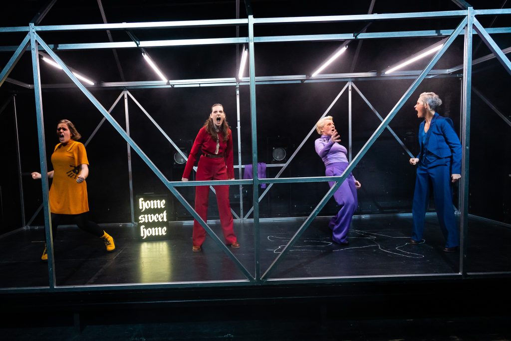 Inside a cube made of aluminum struts, four performers in different colored costumes scream. Between them stands a neon sign with the words “Home Sweet Home.”