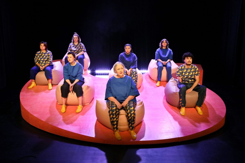 Six performers sit on pink beanbags arranged in a staggered formation on a geometrically shaped, sloping pink platform. Their blue costumes are similar. At the front of the formation is a comparatively older performer who speaks thoughtfully to the audience.