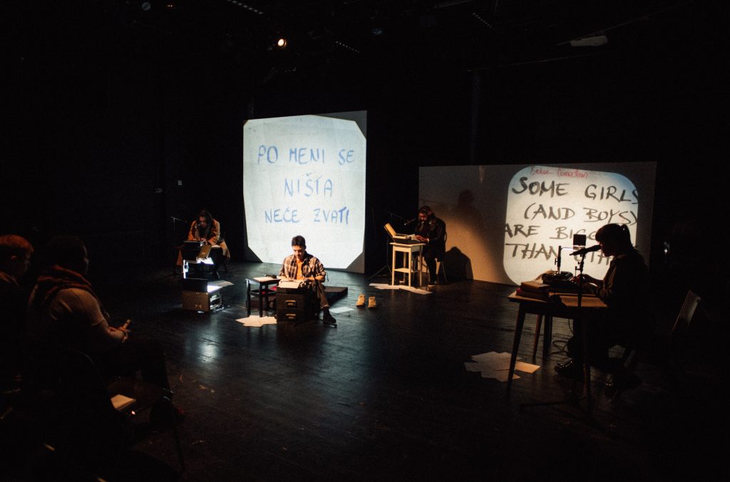 Eine Vorstellung im Rahmen des South East Festival am Theater Oberhausen: auf der abgedunkelten Bühne sitzen vier Menschen vereinzelt - an einem Schreibtisch, vor einem Hocker, vor einem Computer. Sie lesen Text ab, der zugleich zweisprachig im Hintergrund auf die Wand projiziert wird.