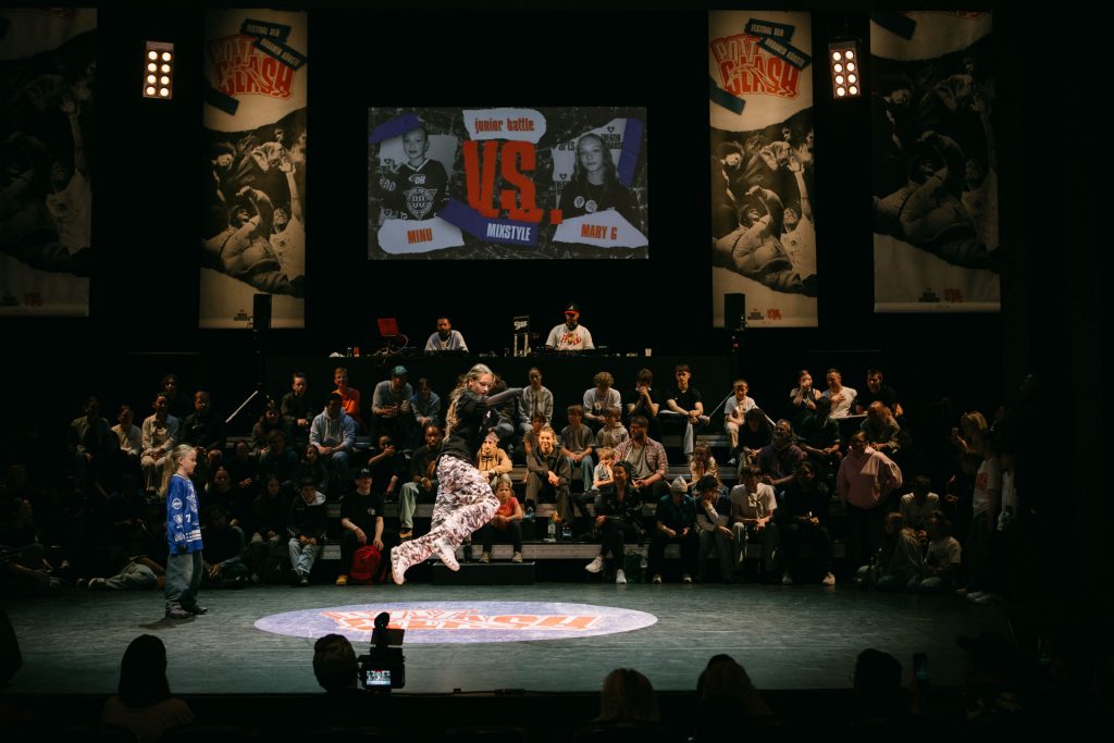 Scene from the Kids Battle at the PottClash Festival at Theater Oberhausen: a young dancer floats in midair in the middle of a movement. In the background, other young people sit on a rising grandstand. A DJ booth is located in front of large banners announcing the respective battle.