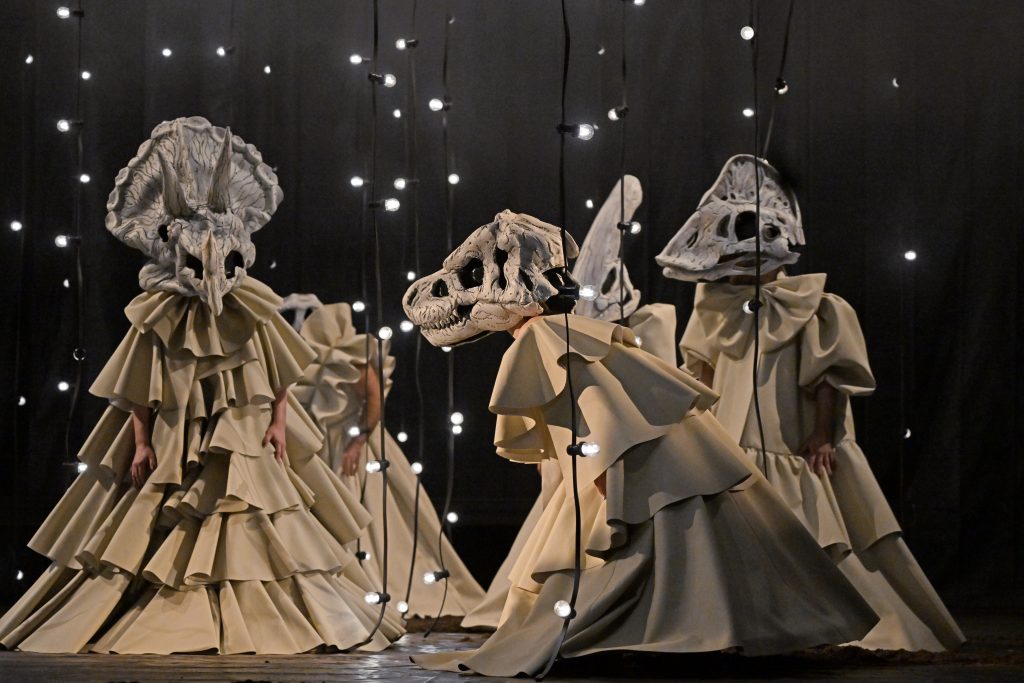 Between large strings of lights hanging from the ceiling, four performers move around in impressive costumes: flowing, puffy beige dresses conceal their bodies, and they wear dinosaur masks on their heads.