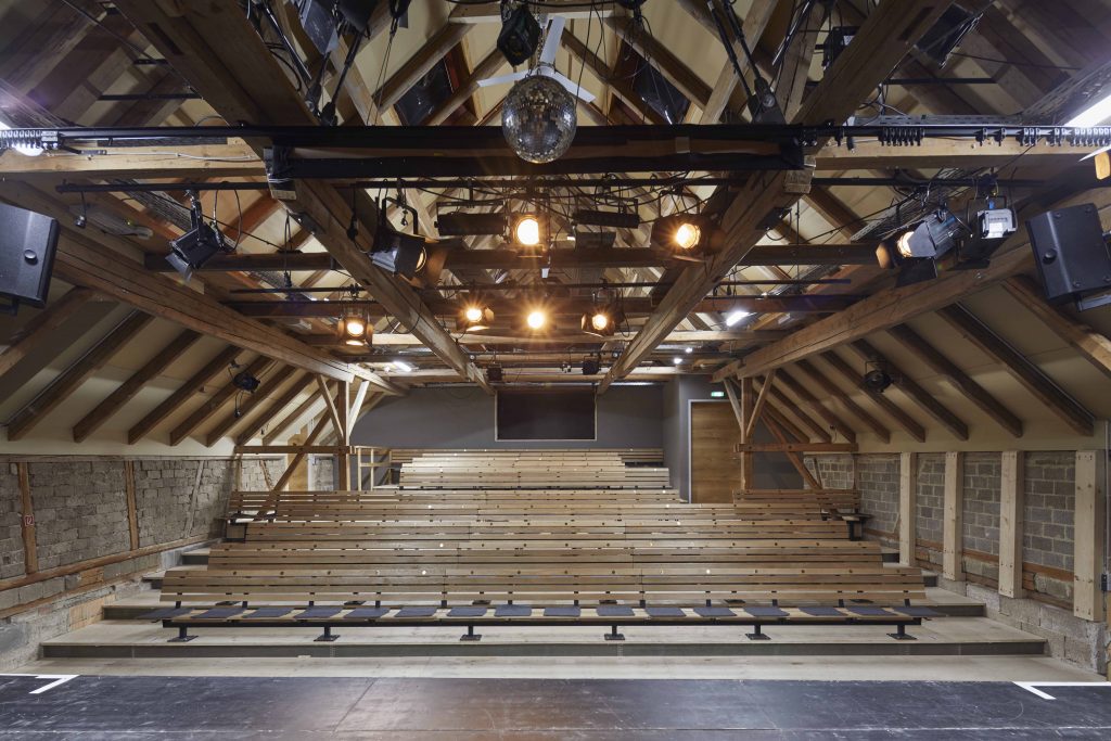 View of the auditorium at Theater Lindenhof: a disco ball hangs beneath the open roof trusses. In front of the ground-level stage is a grandstand with seating for the audience.