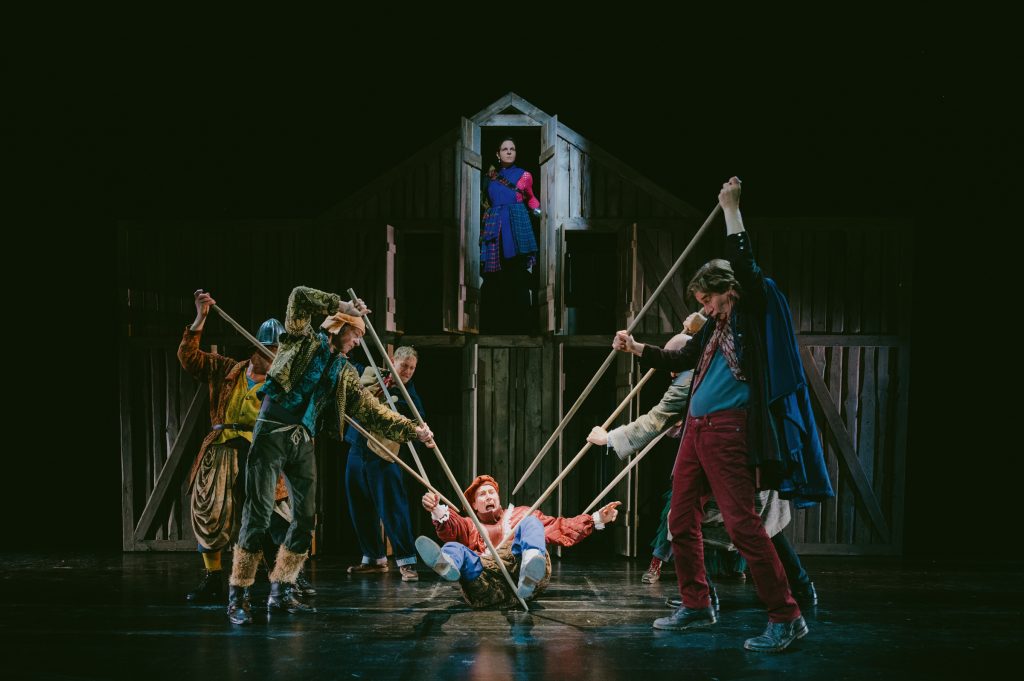 Six people in old-fashioned costumes threaten a person lying on the ground with long sticks, who appears to be screaming loudly. In the background, a wooden house can be seen, with a woman looking down on the commotion from the upper floor.