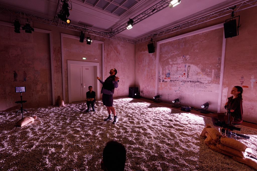Scene from “THE GREAT GRAND OTHER”: a man stands on stage with his hands raised, shouting. Two other people sit on the left and right, watching him. The floor is covered with gravel, and the stage is bathed in pink light.