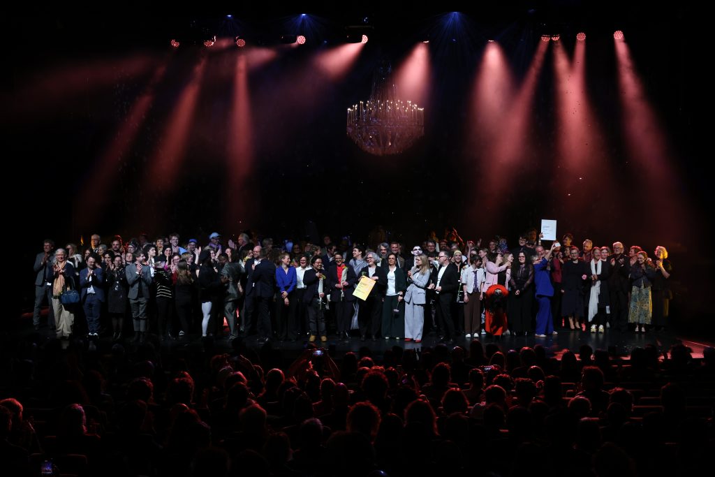 Bühnensituation mit Kronleuchter und farbigem Lichtspiel. Auf der Bühne Preisträger*innen und Programmbeteiligte. Vor ihnen das Publikum.