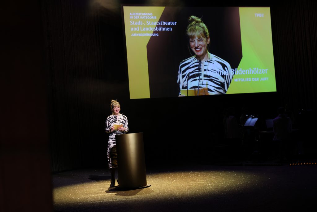 Yvonne Büdenhölzer steht am goldenen Pult und schaut lachend ins Publikum. Hinter ihr eine Projektion von ihr mit ihrem Namen und den Hinweis auf die Preiskategorie.