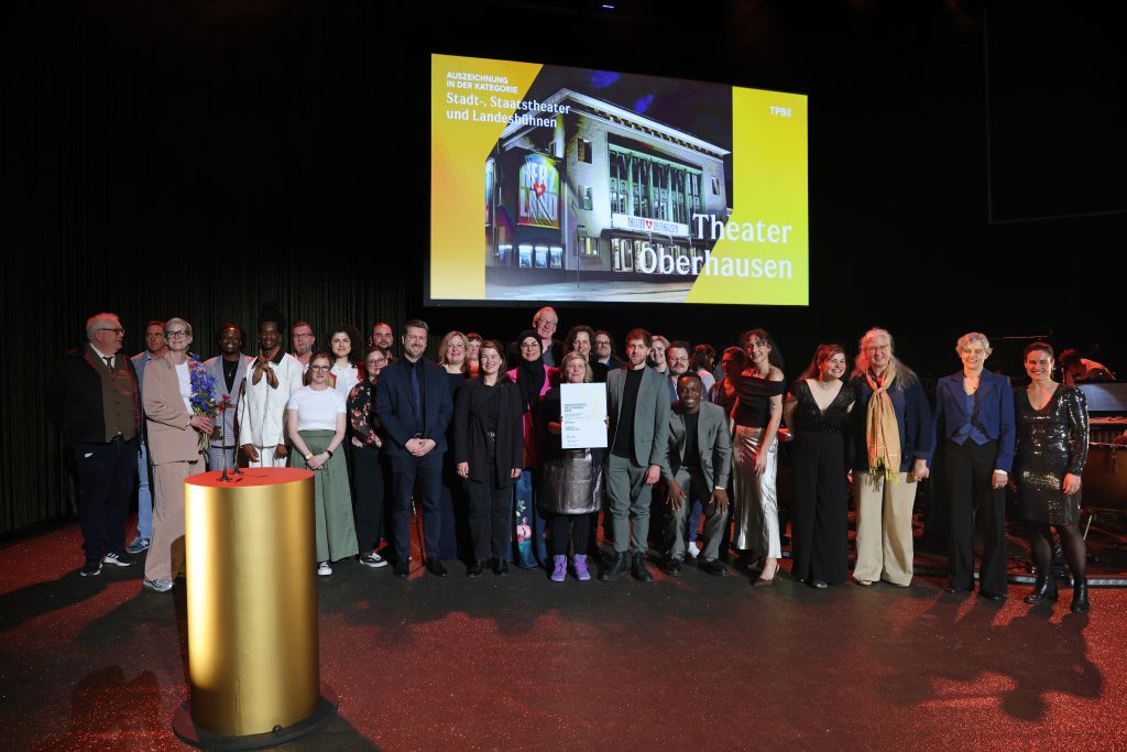 Die Mitarbeiter*innen des Theater Oberhausen stehen auf der Bühne, halten Blumen und eine Urkunde. Hinter ihnen eine Projektion mit des Außenansicht des Theaters.