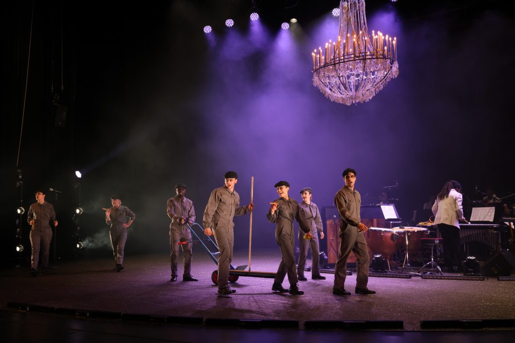 Schauspieler*innen sind auf der Bühne, tragen graue Overalls, eine Schiebermütze und halten Zigarren in den Händen.