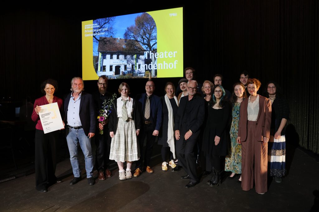 Mitarbeiter*innen des Theater Lindenhof halten Blumen und Urkunden. Hinter ihnen eine Projektion mit der Außenansicht des Theaters.