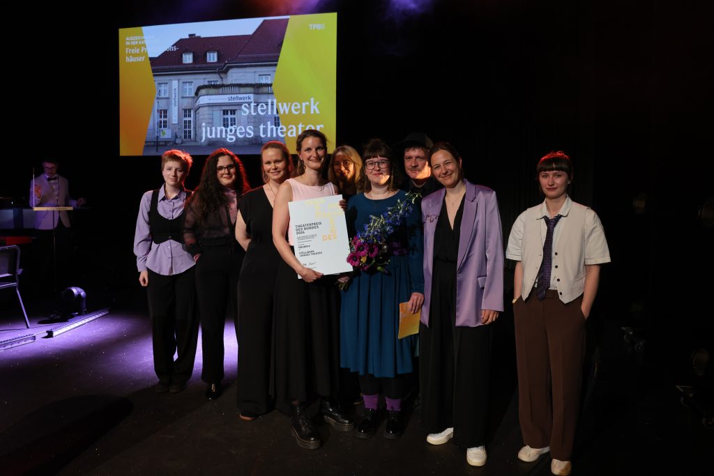 Das Team des stellwerks hält Blumen und Urkunden. Hinter ihnen eine Projektion mit der Außenansicht des Theaters.