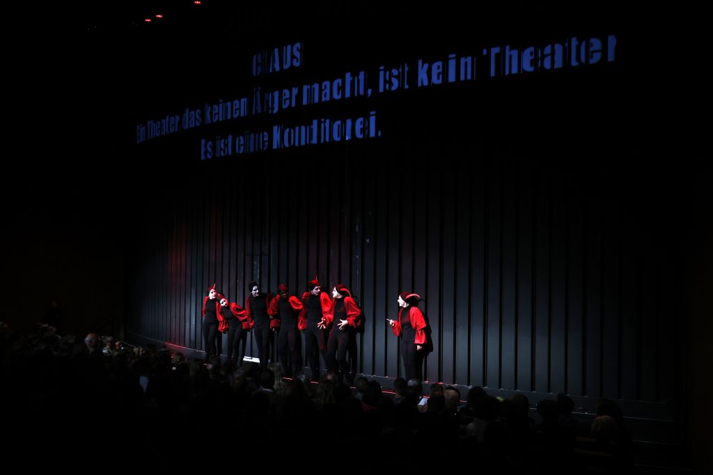 7 Schauspieler*innen tragen ein Mephisto-Kostüm. Hinter ihnen der Eiserne Vorhang auf den ein Zitat projiziert wird.