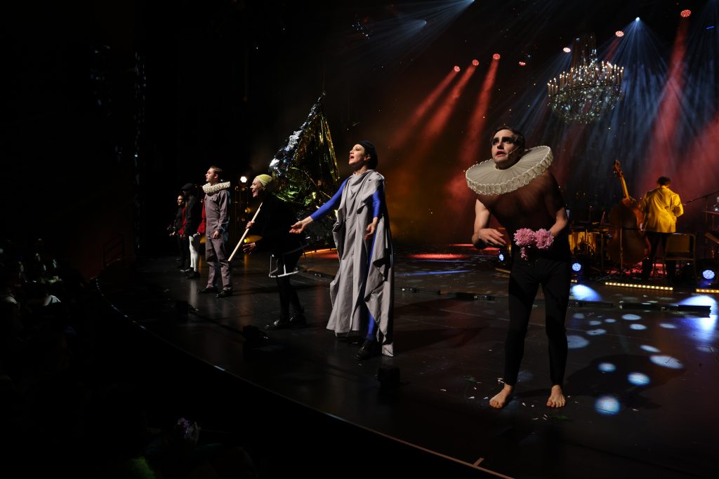 Schauspieler*innen stehen an der Rampe und haben verschiedene Kostüme aus den vorangegangen Szenen an. Eine Schauspielerin hält eine große goldene Fahne.