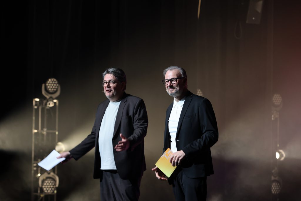 Matthias Pees und Holger Bergmann stehen nebeneinander. Beide tragen einen dunklen Anzug und halten Moderationskarten vor sich.