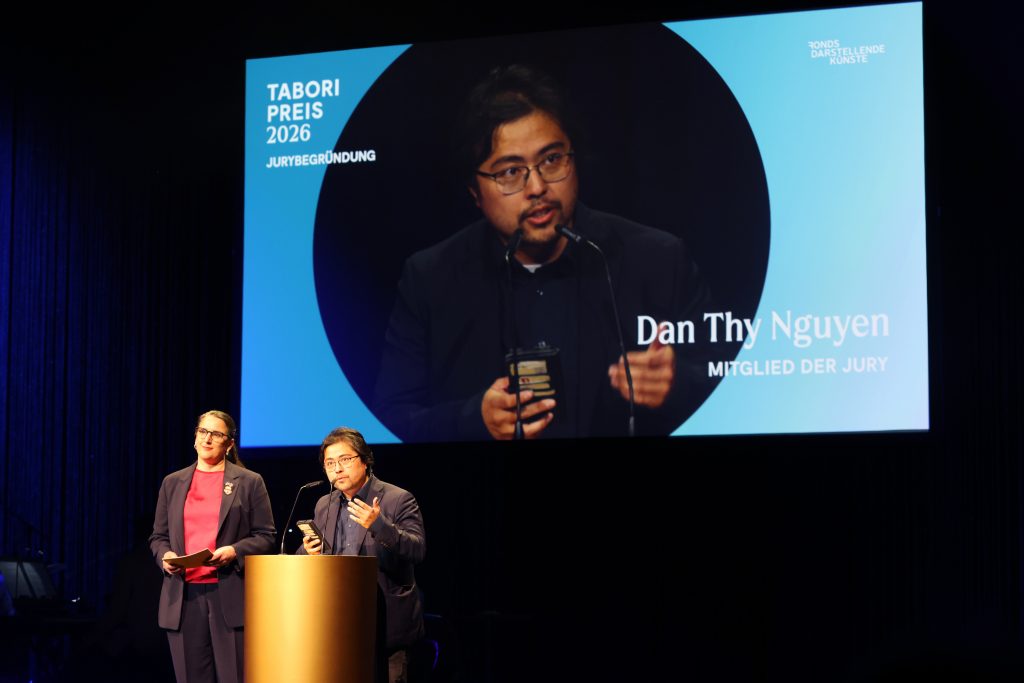 Beide Juror*innen stehen am Pult. Auf der Projektion im Hintergrund ist Dan Thy Nguyen.