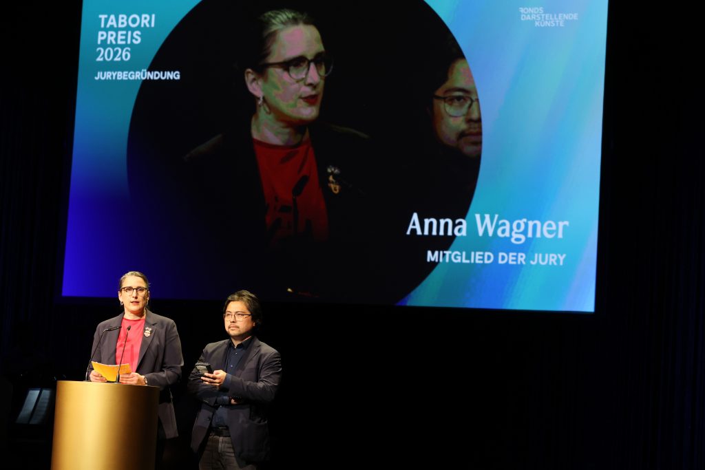 Beide Juror*innen stehen am Pult auf der Projektion im Hintergrund ist Anna Wagner zu sehen.