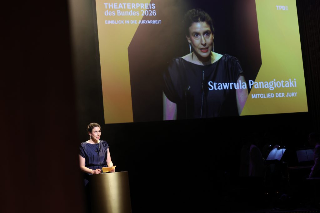 Stawrula Panagiotaki steht am goldenen Pult. Sie schaut ins Publikum. Hinter ihr ist eine Projektion von ihr mit ihrem Namen und dem Zusatz, dass sie Teil der Jury 2026 war.