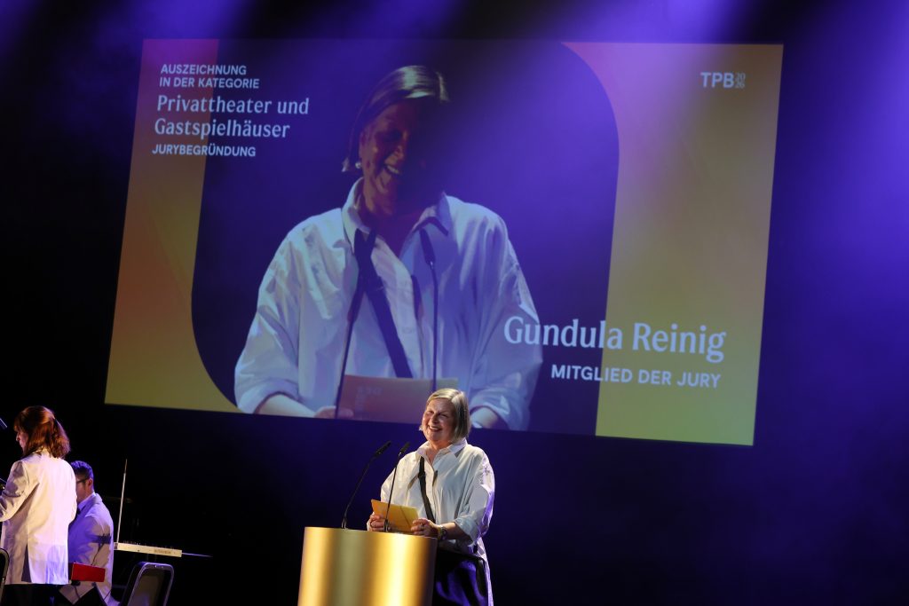 Gundula Reinig steht am goldenen Pult. Sie hält Moderationskarten und schaut ins Publikum. Hinter ihr eine Projetkion von ihr mit ihrem Namen und dem Hinweis auf die Preiskategorie.