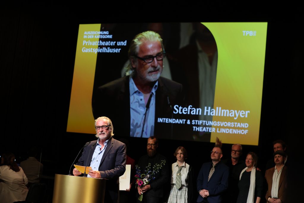 Intendant Stefan Hallmayer steht am Pult, hinter ihm stehen Mitarbeiter*innen des Theaters. Im Hintergrund eine Projektion von Hallmayer.
