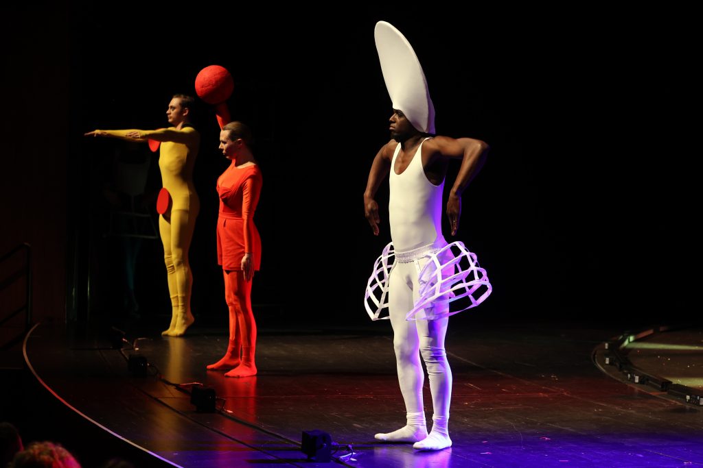 Drei Schauspieler*innen auf der Bühne in einem gelben, roten und weißem Kostüm an denen Kreise und Kugeln angebracht sind.