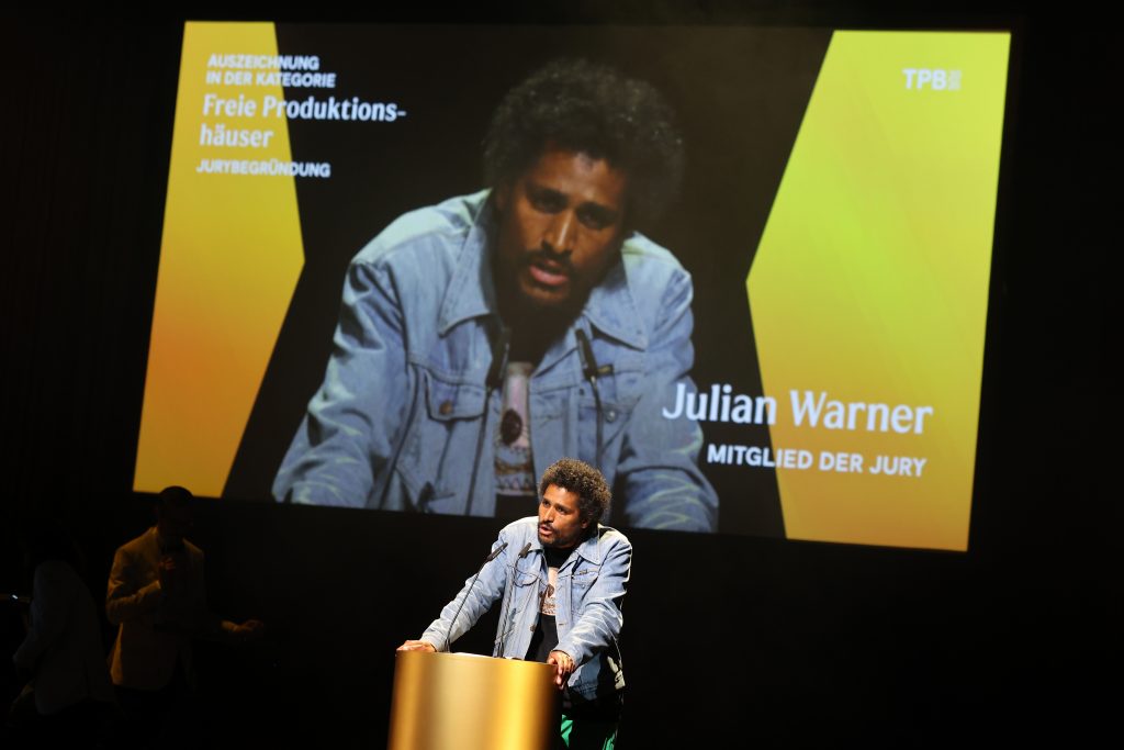 Julian Warner steht am Pult und schaut ins Publikum. Hinter ihm eine Projektion von ihm mit seinem Namen und dem Hinweis auf die Preiskategorie.