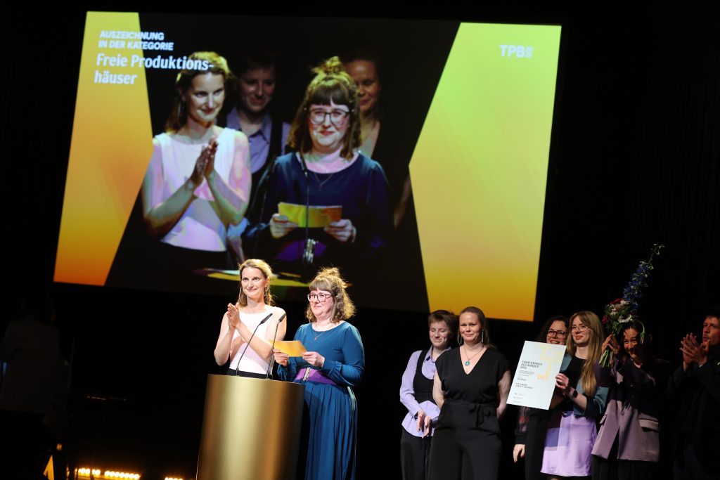 Stefanie Heiner und Juliane Heinrich stehen am Pult ihr Team hinter ihnen. Projektionen von den beiden auf der Leinwand im Hintergrund.