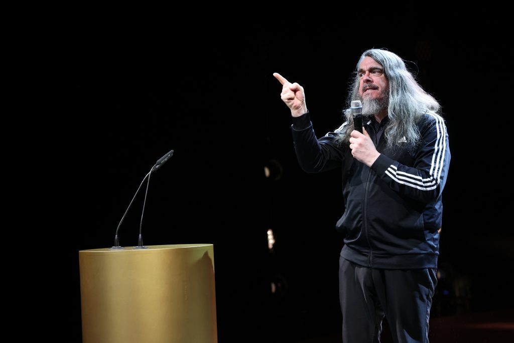 Jonathan Meese spricht in ein Handmikrofon und hat die rechte Hand mit erhobenem Zeigefinger nach vorn ausgestreckt.