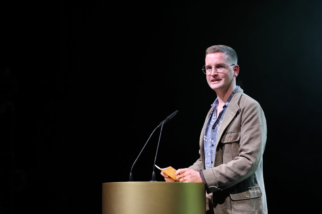 Clemens Leander steht an einem goldenen Pult mit Mikorofon und hält Moderationskarten vor sich. Er schaut ins Publikum.