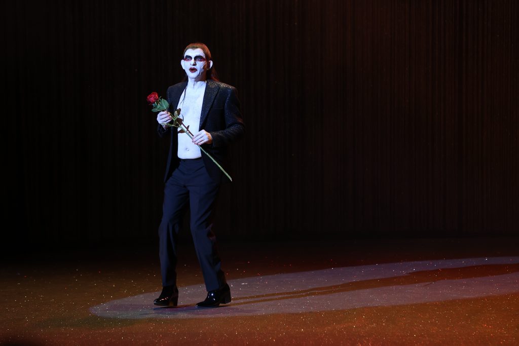 Dominik Horwitz hat ein weiß geschminktes Gesicht und trägt eine dunkle Perücke. Sein Jacket ist offen, darunter der freie Oberkörper. Er hält vor sich eine rote Rose.